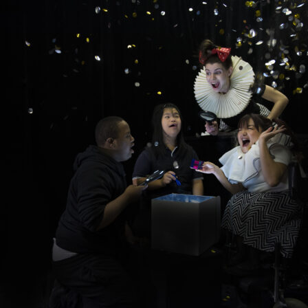 Two female performers, including one in a wheelchair, and two children with Down syndrome are peeping into a magical cardboard box with magnificent glasses. They are filled with joy and excitement as a shower of gold and silver shiny confetti fall on top of them.
