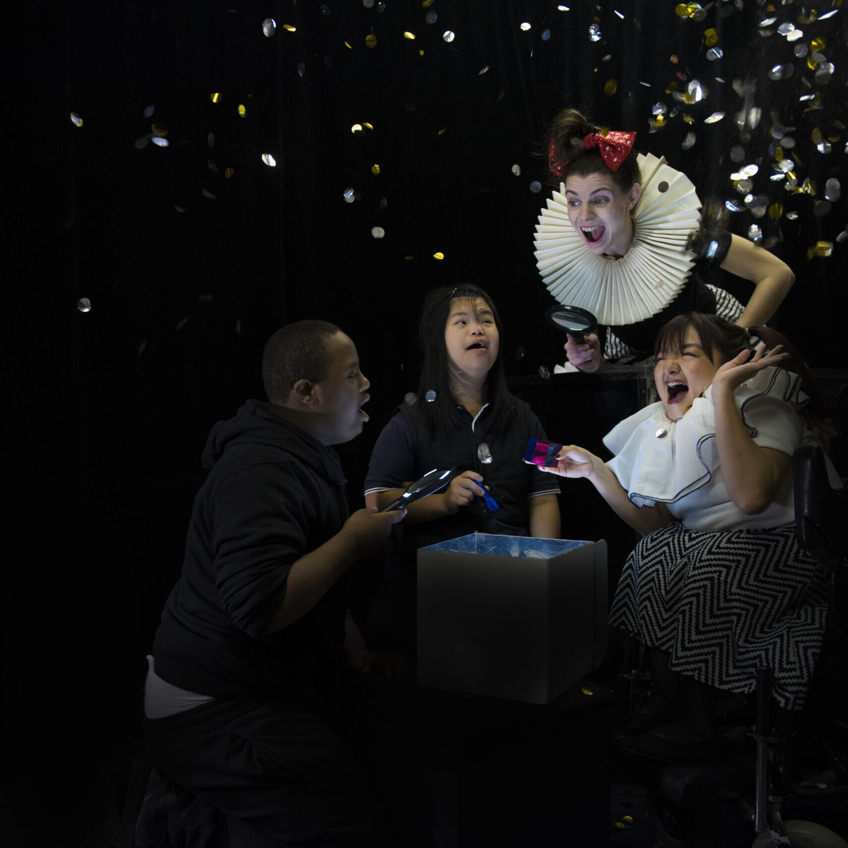 Two female performers, including one in a wheelchair, and two children with Down syndrome are peeping into a magical cardboard box with magnificent glasses. They are filled with joy and excitement as a shower of gold and silver shiny confetti fall on top of them.